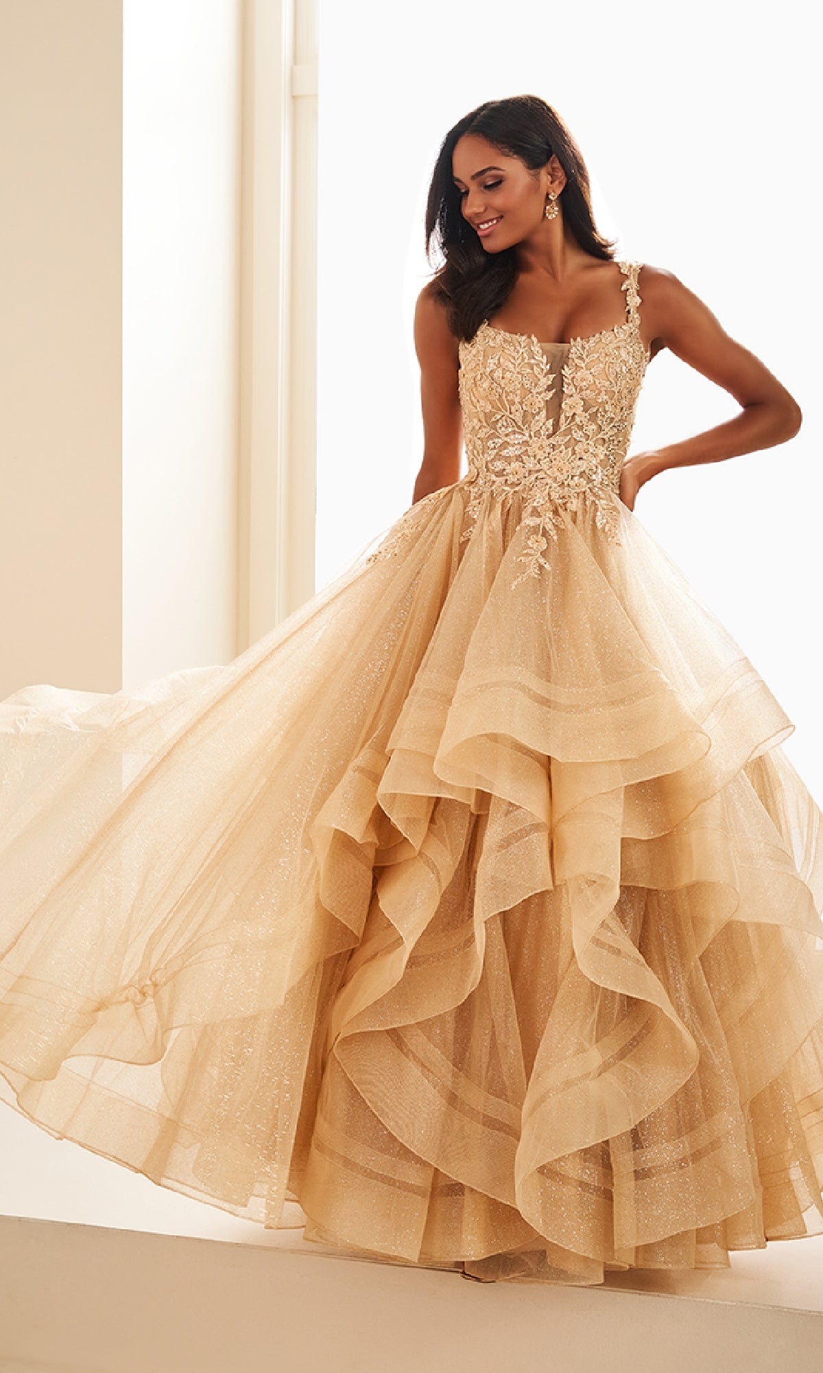 A woman stands indoors by a bright window, holding the skirt of the Ellie Wilde EW36035 Long Glitter Prom Ball Gown featuring a layered gold design and sequin-lace bodice.